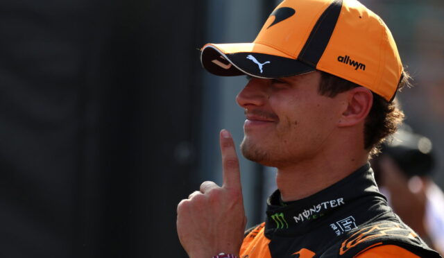 Portrait of a male race driver in an orange cap and racing suit, with his finger to his lips in a shushing gesture.
