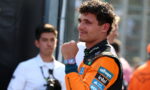 Racing driver in orange suit adjusts his wristwatch, looking to the side, with sponsor patches on the suit and a blurred crew member in the background.