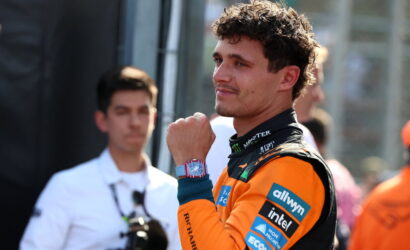 Racing driver in orange suit adjusts his wristwatch, looking to the side, with sponsor patches on the suit and a blurred crew member in the background.