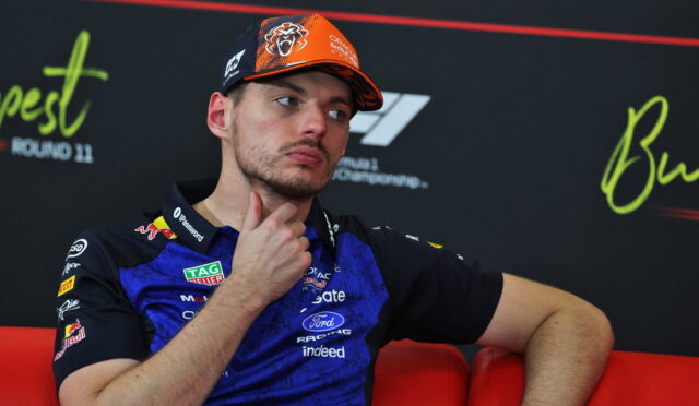 Male race car driver seated on a red couch during a press event, wearing a blue team polo and orange cap, hand on chin, looking to the side.