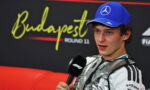 Driver in a silver team suit and blue cap speaks into a microphone during a post‑race interview at Budapest Round 11.