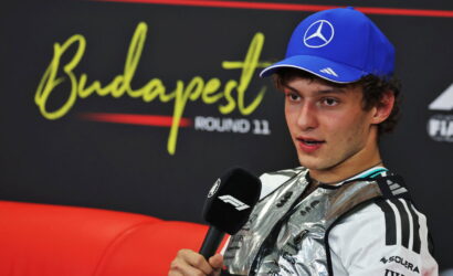 Driver in a silver team suit and blue cap speaks into a microphone during a post‑race interview at Budapest Round 11.