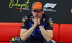 F1 driver in Oracle Red Bull Racing gear sits on a red couch with hands clasped under the chin, looking thoughtful.
