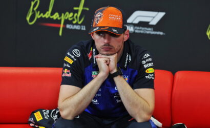 F1 driver in Oracle Red Bull Racing gear sits on a red couch with hands clasped under the chin, looking thoughtful.