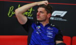 F1 driver in a blue racing shirt sits on a red couch, hand on head, looking thoughtful during an interview backdrop with sponsor logos.