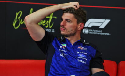 F1 driver in a blue racing shirt sits on a red couch, hand on head, looking thoughtful during an interview backdrop with sponsor logos.