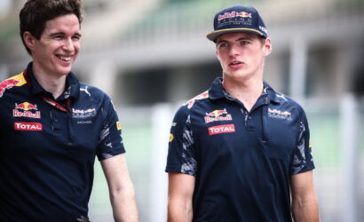 Two men in Red Bull Racing polo shirts walk and chat at a race track, smiling.