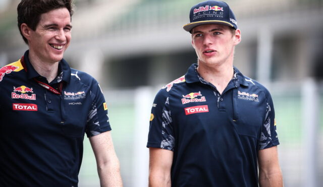 Two men in Red Bull Racing polo shirts walk and chat at a race track, smiling.