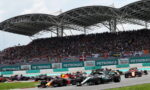 Formula 1 cars race off the start on a track, with a packed grandstand beneath a curved steel canopy.