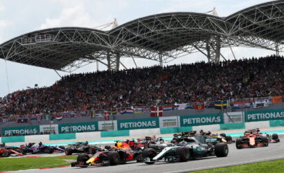 Formula 1 cars race off the start on a track, with a packed grandstand beneath a curved steel canopy.