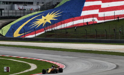 Yellow Formula 1 car rounds a curve on a race track with a large Malaysian flag mural on the hill behind.