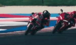 Two red racing motorcycles lean into a corner on a racetrack, riders in red gear close together.