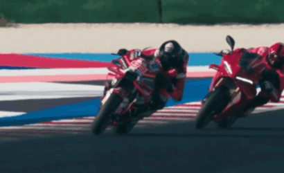 Two red racing motorcycles lean into a corner on a racetrack, riders in red gear close together.