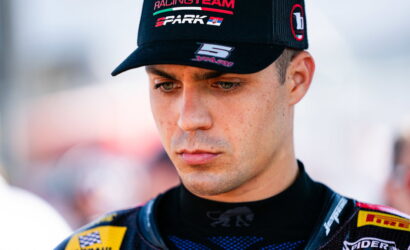 Close-up of a male racing driver wearing a black cap, focused, in a sponsor-branded race suit.