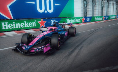 Formula 1 car in pink and blue livery speeding along a street circuit with Heineken banners in the background