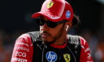 Portrait of a Formula 1 driver wearing a red Ferrari cap, dark sunglasses, and earphones, looking to the side with sponsor logos on his suit.