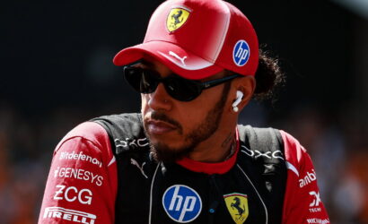 Portrait of a Formula 1 driver wearing a red Ferrari cap, dark sunglasses, and earphones, looking to the side with sponsor logos on his suit.