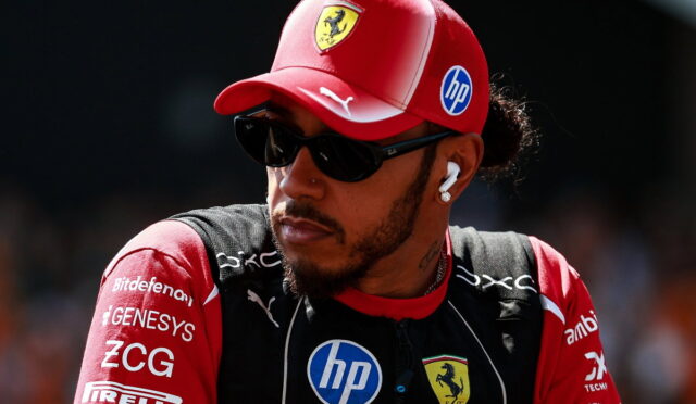 Portrait of a Formula 1 driver wearing a red Ferrari cap, dark sunglasses, and earphones, looking to the side with sponsor logos on his suit.