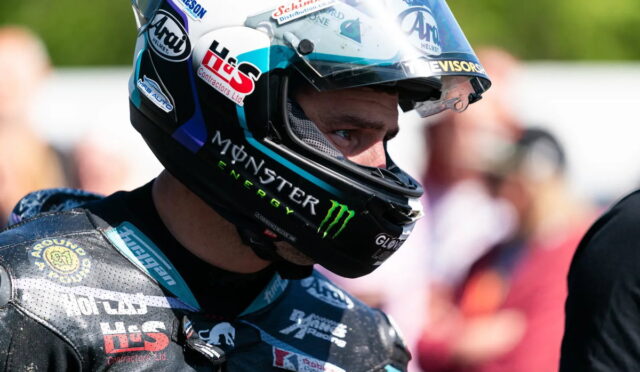 Motorcycle racer in a full-face helmet with Monster Energy and sponsor logos, ready at the track.