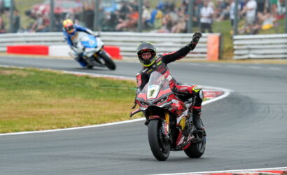 Moto racing rider on a red Ducati leads into a bend, raising a gloved fist in celebration as a blue bike tracks behind with spectators in the background.