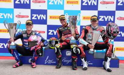 Three motorcycle racers on a winners' podium hold large trophies, smiling in racing suits with helmets nearby against a sponsor backdrop.