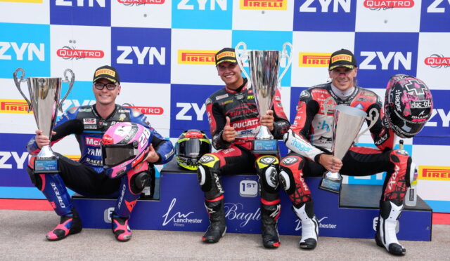 Three motorcycle racers on a winners' podium hold large trophies, smiling in racing suits with helmets nearby against a sponsor backdrop.