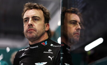 Portrait of a male race car driver in a black Aston Martin racing suit, standing beside a glass barrier with a contemplative expression.