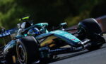 Green Formula 1 race car on a track, close-up side view showing aerodynamic body and sponsor decals in motion on asphalt.