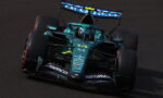 Green Formula 1 race car navigating a turn on a dark racetrack, sponsor logos visible on the nose and wings.