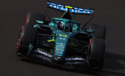 Green Formula 1 race car navigating a turn on a dark racetrack, sponsor logos visible on the nose and wings.