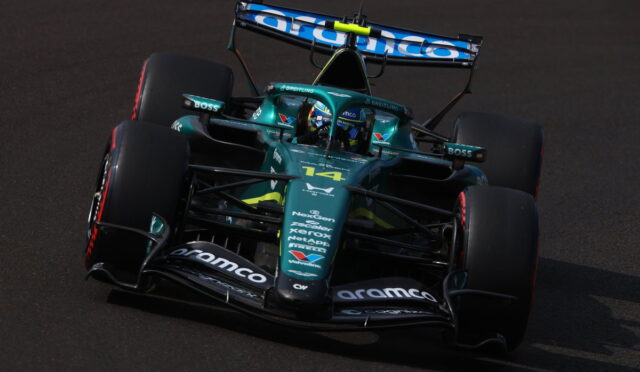 Green Formula 1 race car navigating a turn on a dark racetrack, sponsor logos visible on the nose and wings.