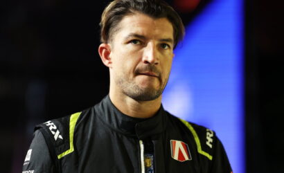 Male race car driver in a black racing suit with neon yellow accents, looking pensively to the side, against a dark backdrop with blue lighting.