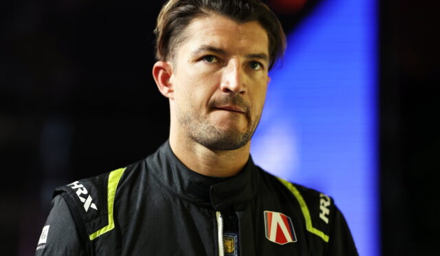 Male race car driver in a black racing suit with neon yellow accents, looking pensively to the side, against a dark backdrop with blue lighting.