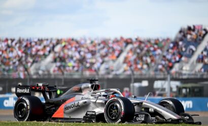 A silver Formula 1 car with Revolut branding speeding on the track, crowd-filled grandstands blurred in the background.