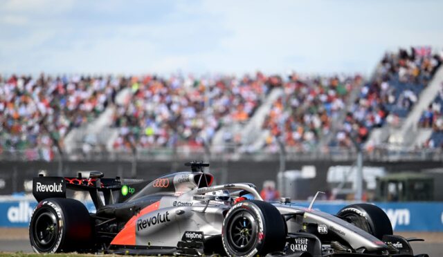 A silver Formula 1 car with Revolut branding speeding on the track, crowd-filled grandstands blurred in the background.