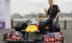 Smiling man in a racing shirt sits on a Red Bull Formula 1 car, giving a peace sign, with a city skyline in the background.