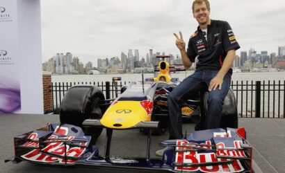 Smiling man in a racing shirt sits on a Red Bull Formula 1 car, giving a peace sign, with a city skyline in the background.