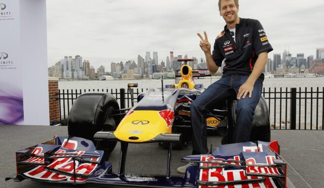 Smiling man in a racing shirt sits on a Red Bull Formula 1 car, giving a peace sign, with a city skyline in the background.