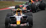 Front-row Formula 1 car with orange nose and Red Bull livery leading a pack on a race track; sponsor logos visible on the car’s nose and wings.