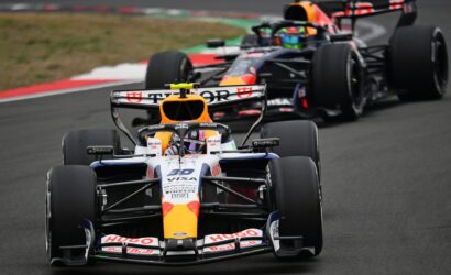 Front-row Formula 1 car with orange nose and Red Bull livery leading a pack on a race track; sponsor logos visible on the car’s nose and wings.
