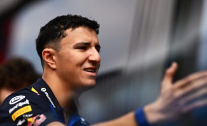 Close-up of a young man in a racing team shirt smiling at an event, with sponsor logos on his sleeve.