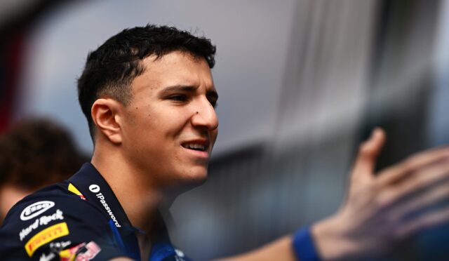 Close-up of a young man in a racing team shirt smiling at an event, with sponsor logos on his sleeve.