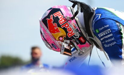 Close-up of a colorful racing helmet with Red Bull branding, attached to a blue racing suit, against a bright sky.
