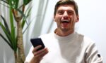 Young man with short brown hair and stubble, laughing while holding a smartphone; a large green plant is in the background.