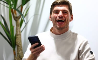 Young man with short brown hair and stubble, laughing while holding a smartphone; a large green plant is in the background.