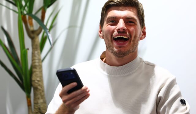 Young man with short brown hair and stubble, laughing while holding a smartphone; a large green plant is in the background.