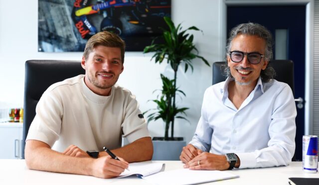 Two men in an office sign documents at a white table, smiling at the camera.
