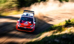 Rally car in red and white livery speeding on a dirt road, kicking up dust as it leans into a bend.