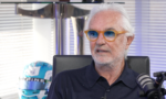 Older man with white hair and orange glasses speaks into a microphone in a studio.