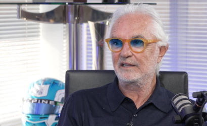 Older man with white hair and orange glasses speaks into a microphone in a studio.